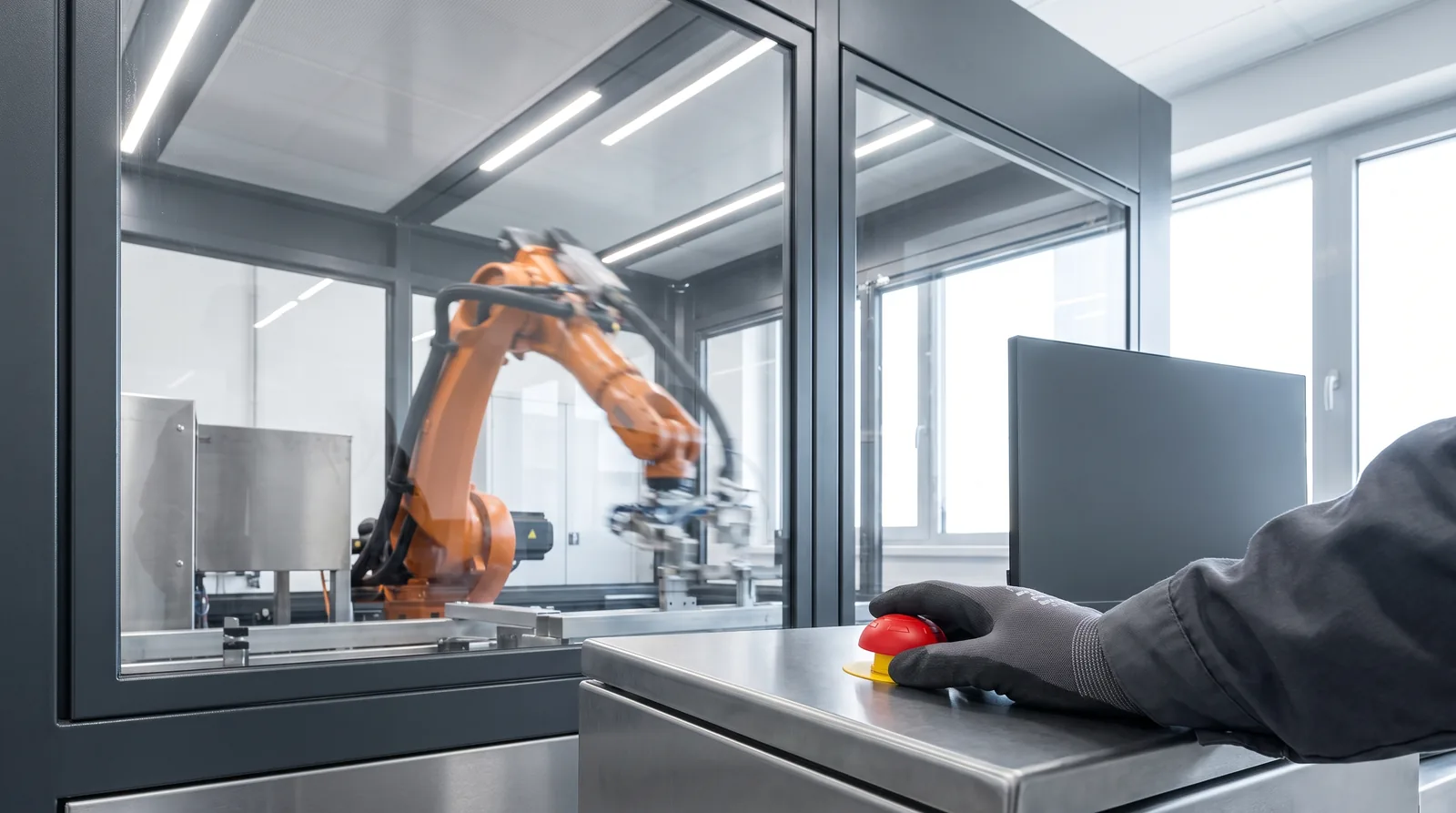 Dłoń operatorki opiera się obok czerwonego przycisku zatrzymania awaryjnego na pulpicie linii produkcyjnej, za szybą pracuje ramię robota